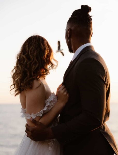 A close-up of a couple from behind, against a serene ocean backdrop during sunset. The man is wearing a dark suit with his hair tied back, embracing a woman in a white off-the-shoulder dress with floral details, whose curly hair cascades down her back.