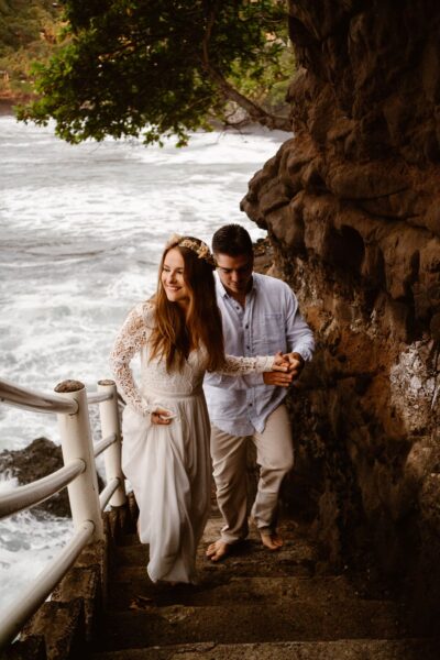 A smiling couple in casual wedding attire descends a stone staircase by the sea, the woman in a white dress and floral headpiece, and the man in a white shirt and beige pants, both barefoot.
