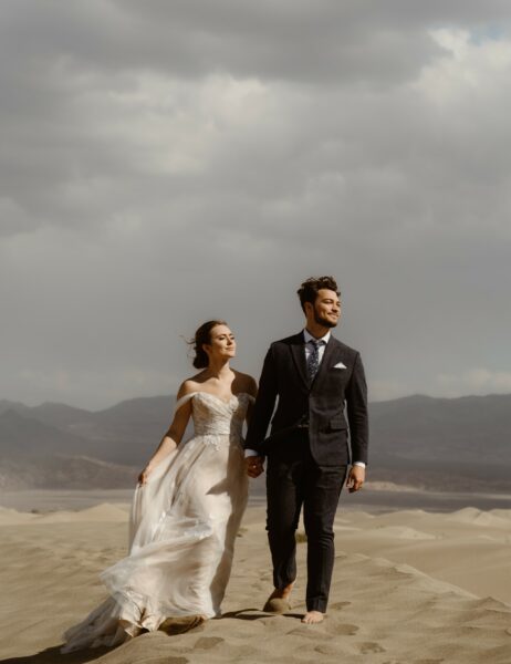 A couple in wedding attire walks hand-in-hand across a desert dune landscape, the woman in a flowing white gown and the man in a tailored suit, both barefoot, with mountains and dramatic clouds in the background.
