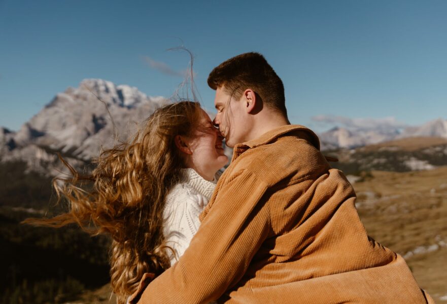 A couple in a joyful embrace, with windswept hair, sharing a loving gaze against the stunning backdrop of the snow-capped Dolomites.