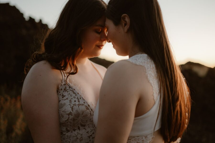 Two women in white dresses share a tender moment, foreheads touching affectionately, as the warm glow of sunset envelops them.