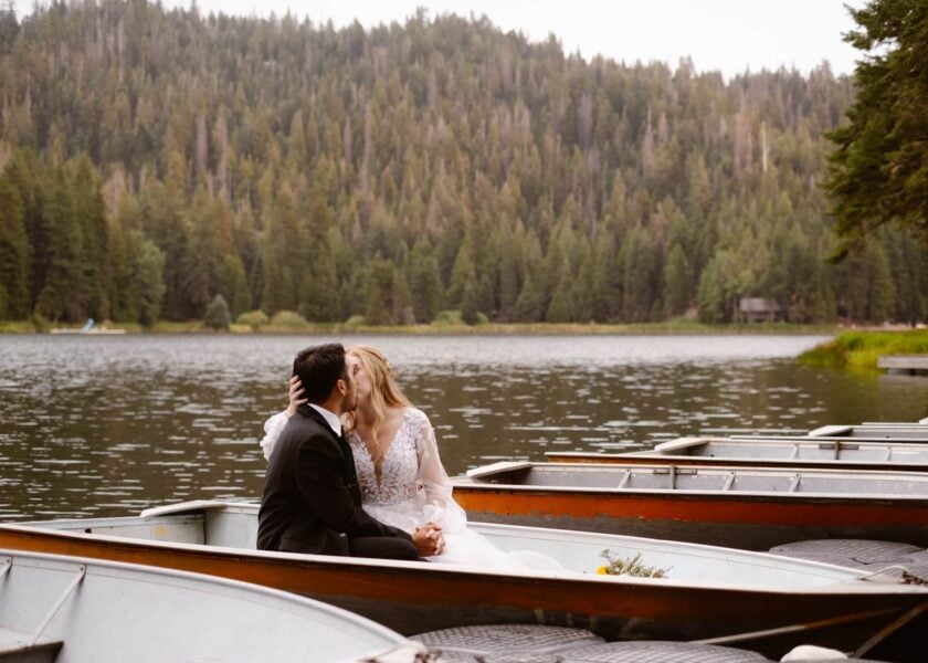 A couple shares a romantic kiss in a rowboat on a tranquil forest lake, surrounded by lush pine trees and the serenity of nature.