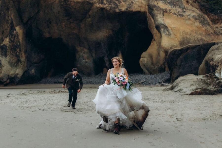Bride running on the sand at Cannon Beach on the Oregon Coast with groom chasing her, dramatic coastal cliffs in the background