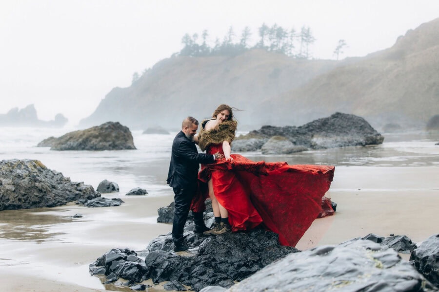 Bride in a flowing red dress standing on rocks with groom at Crescent Beach in Ecola State Park on the Oregon Coast