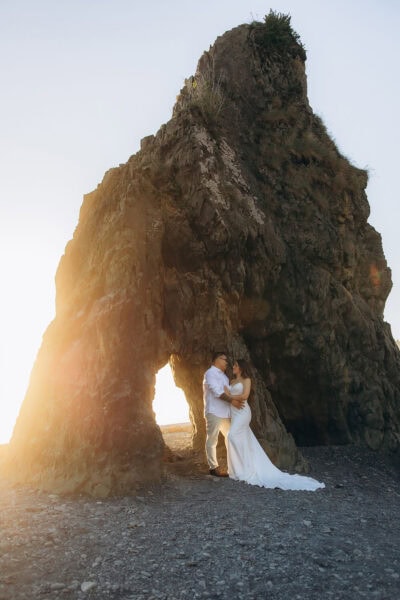 Couple standing beneath Hole in the Wall rock formation at Ruby Beach in Washington during golden hour