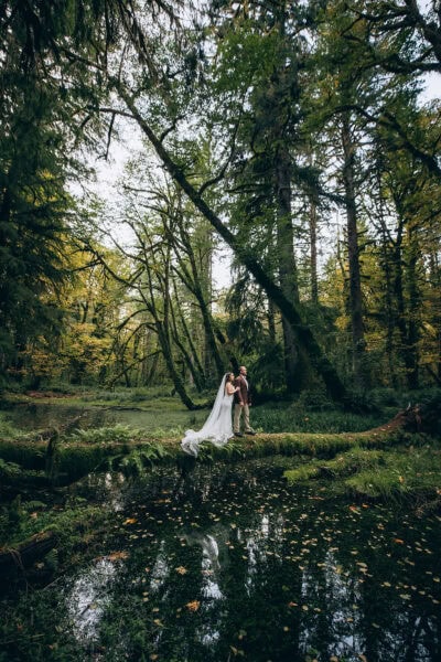 Bride and groom standing on a moss-covered log during a forest elopement in Snoqualmie Valley, Washington, with trees reflected in the water