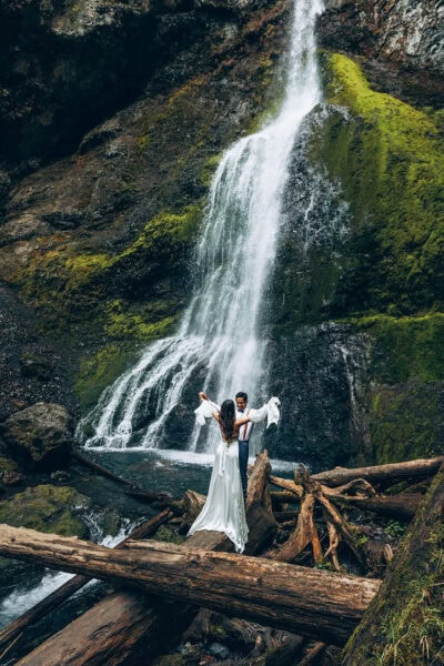 Bride and groom standing on logs at Marymere Falls in Olympic National Park with waterfall cascading behind them