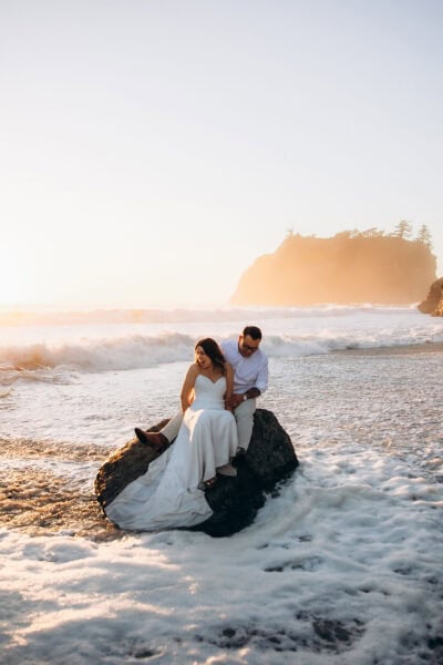 Bride and groom sitting on a rock during sunset on a beach in Olympic National Park with waves washing around them