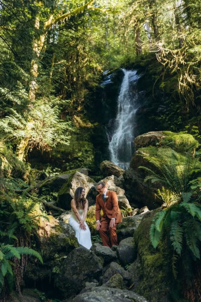 Couple sitting on mossy rocks in front of Merriman Falls in the Quinault Rainforest, Olympic National Park