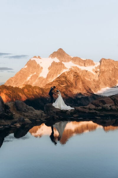 Couple standing on rocks at Huntoon Point with Mount Shuksan glowing at sunset and reflecting in an alpine tarn near Artist Point, Washington