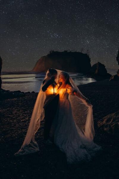 Couple standing under a veil holding a lantern at night on Ruby Beach in Olympic National Park with a star-filled sky above them