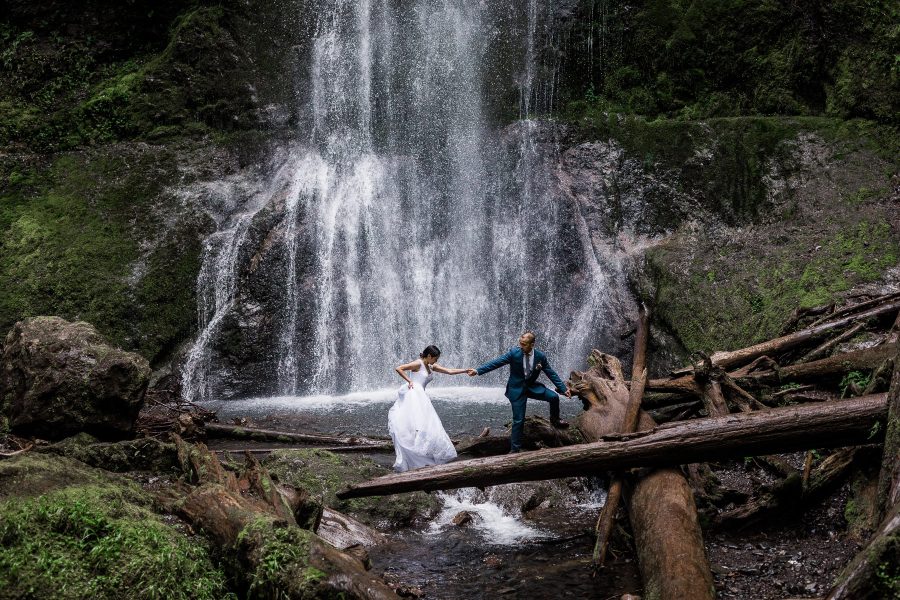 Groom leading a bride across a log in front of a waterfall