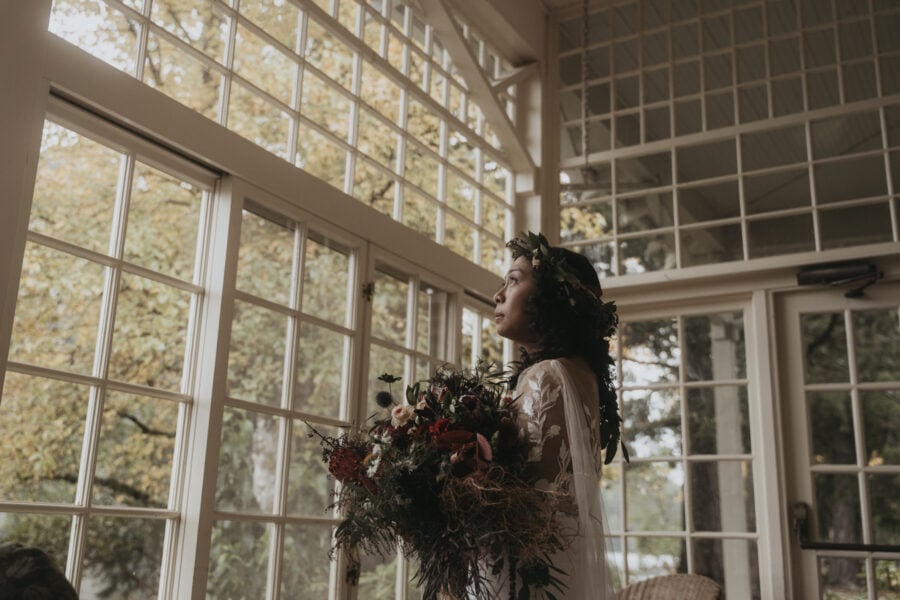 A bride holds her bouquet inside a greenhouse style building, looking out the windows to the lakeside views beyond.
