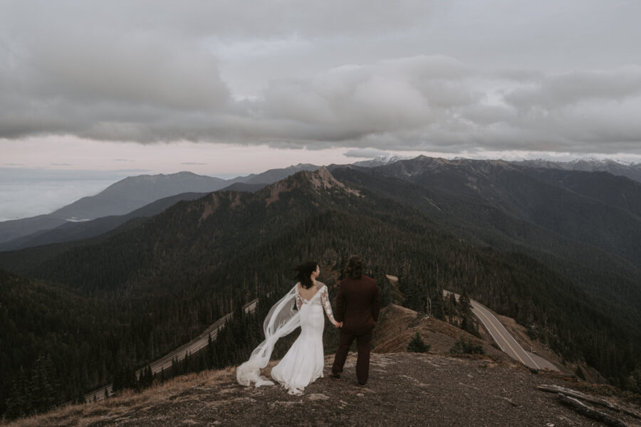 A bride and groom stand facing away from the camera with a road and mountains in the distance.