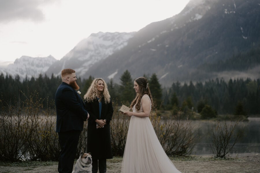 A couple exchanges vows while their bulldog looks on in front of mountains at sunrise.