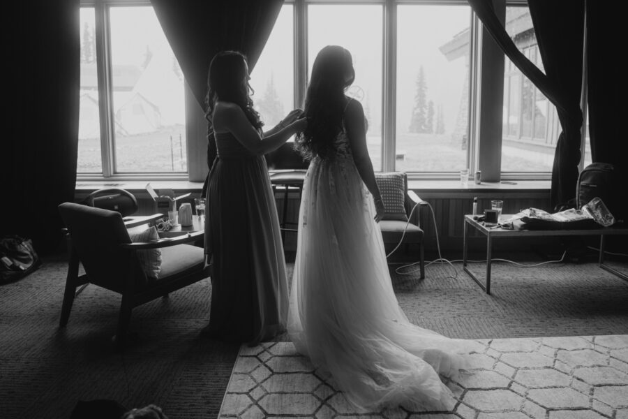 Black and white photo of Jocelyn getting ready for her wedding. Her mom zips up the back of the dress in a lodge with bright windows.