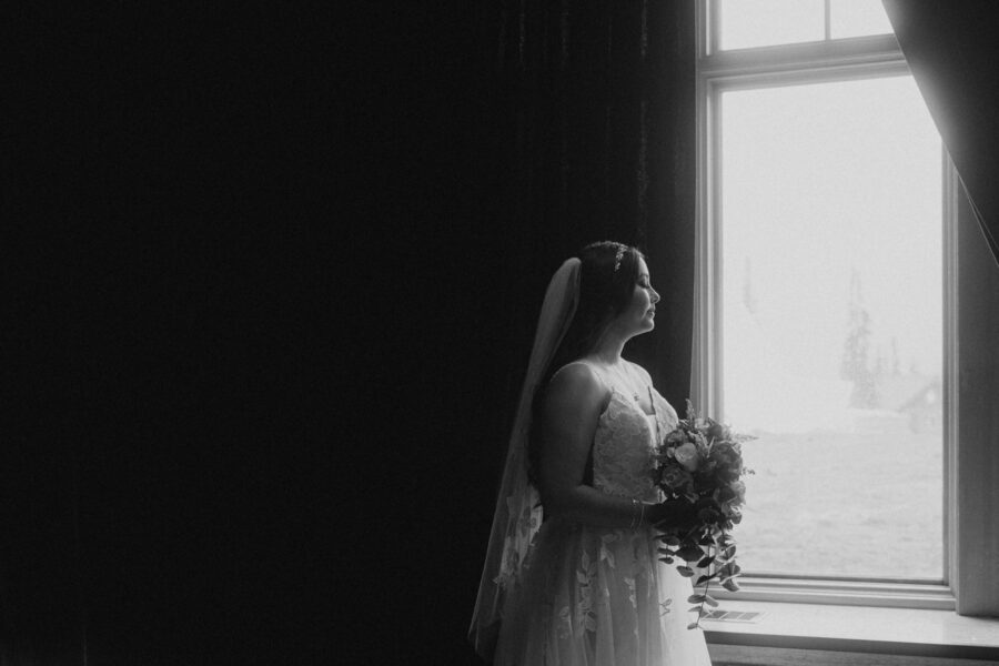 Black and white portrait of a bride holding a bouquet, looking out a window with a view of the mountains.