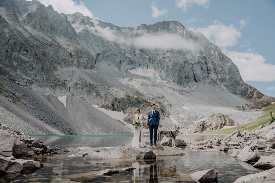 bride and groom and their dog standing on a rock in the middle of an alpine lake
