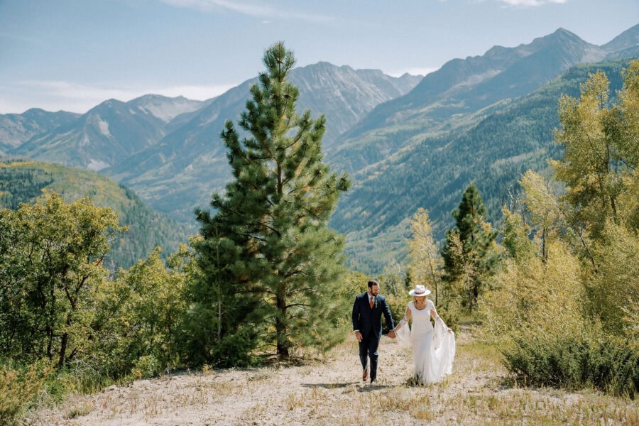 A couple in wedding attire walks hand-in-hand through a forested mountain landscape with tall aspen trees and distant peaks under a clear sky, all beautifully captured by their photographer