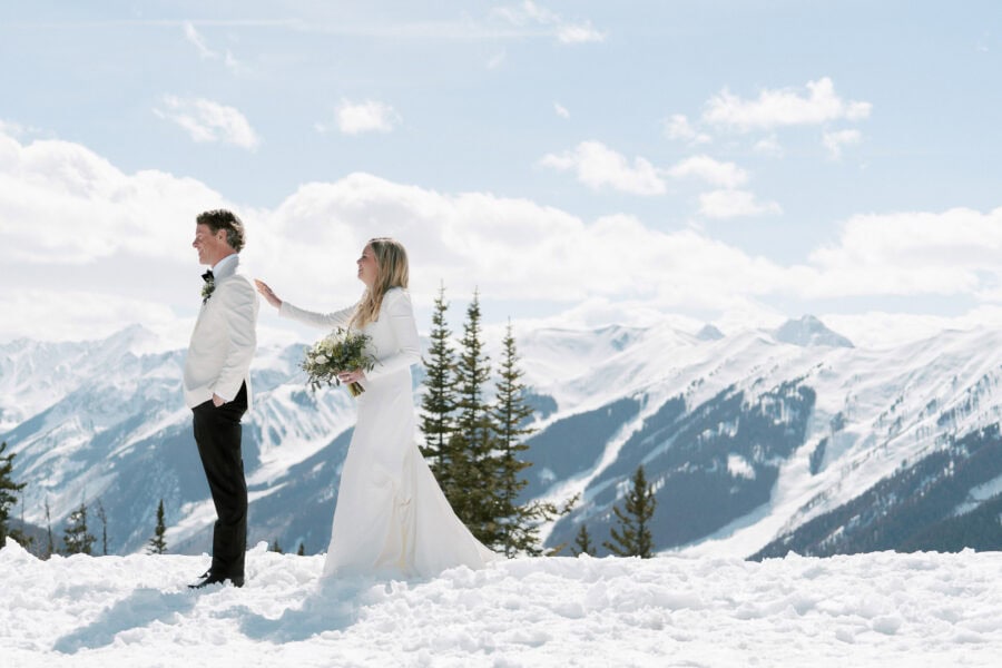 A bride and groom in wedding attire stand in the snow on an Aspen mountain with snow-covered peaks in the background. The bride, holding a bouquet, gently touches the groom's shoulder from behind as he looks forward, creating a serene and romantic scene perfect for any wedding photographer to capture.