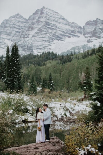 A couple stands embracing on a rock by a serene lake, surrounded by lush greenery and snow-dusted trees. In the background, majestic snow-covered mountains rise under a cloudy sky. The woman holds a bouquet of flowers, while the man wears a light suit. It's an intimate wedding captured in stunning photography.