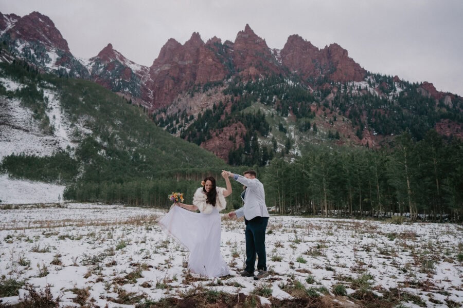 A couple, dressed in wedding attire, dances joyfully in a snowy mountain meadow. The bride wears a long white gown and holds a bouquet, while the groom in a light suit spins her around. Pine trees and rugged, red-tinted mountains create a stunning backdrop.