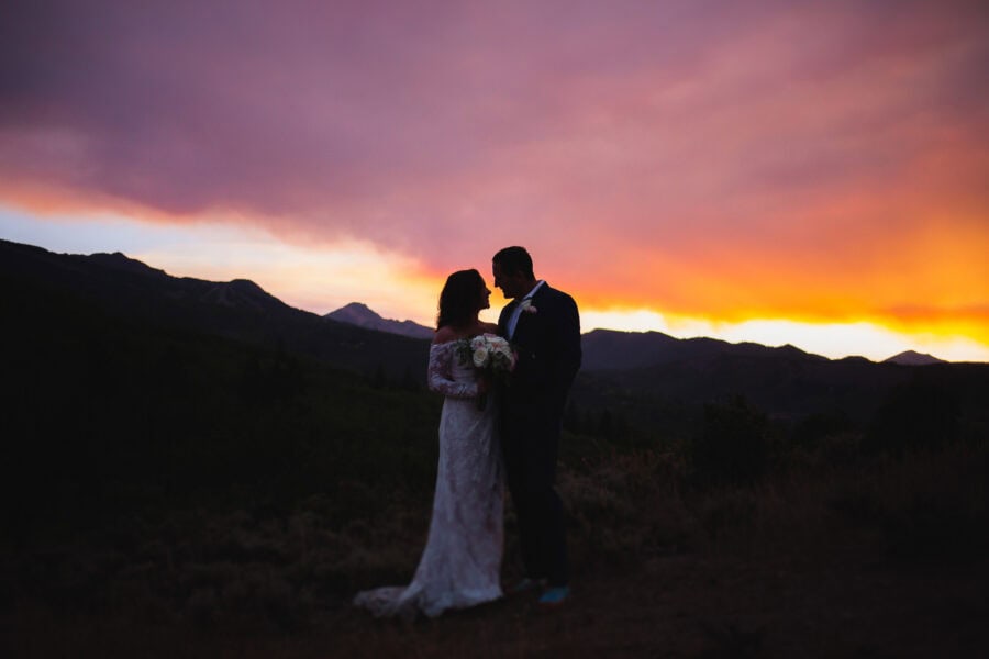 A couple stands close together, holding a bouquet of flowers, against a backdrop of a vibrant sunset with hues of orange, pink, and purple. They are silhouetted against the scenic mountainous landscape. The bride wears a white gown, and the groom is in a dark suit.