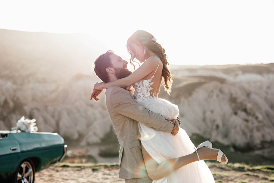 Groom lifting his bride up at sunrise in Cappadocia with blue vintage car behind them