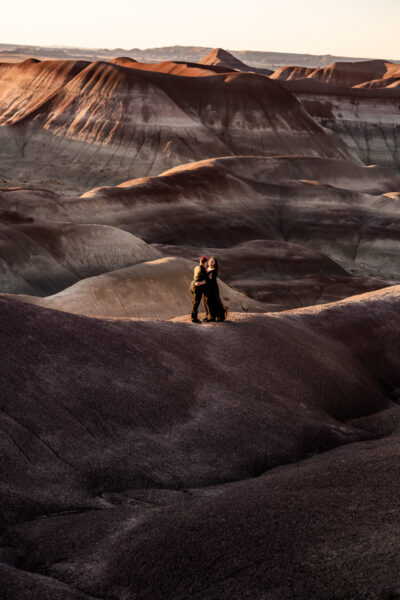 A queer couple stands hand-in-hand in the Painted Desert, one partner in a black wedding dress with a cape.
