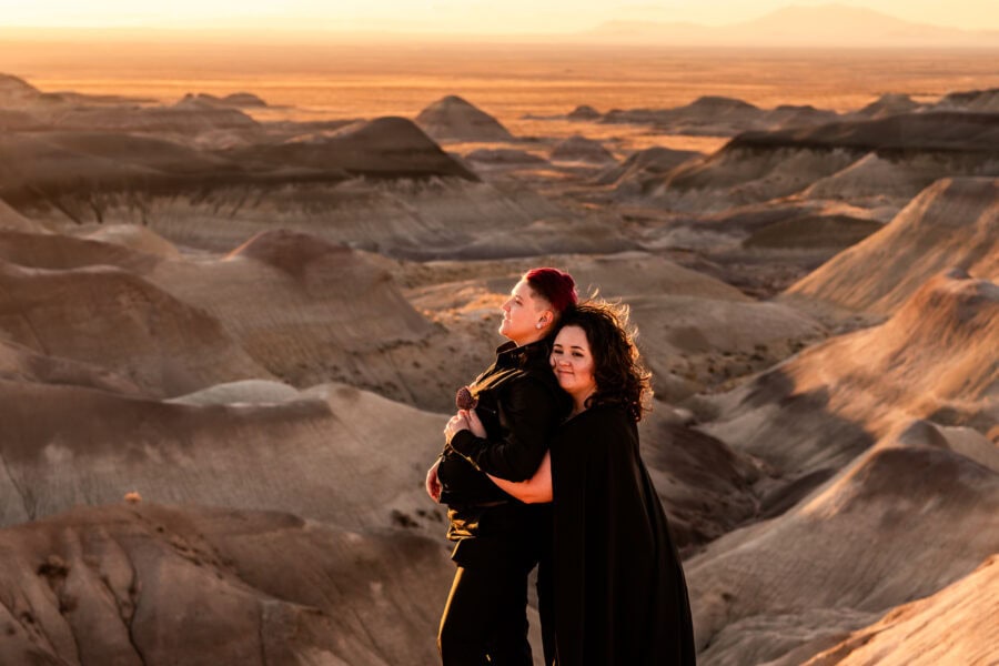 A queer couple in the Painted Desert, one in a black wedding dress with a cape, holding hands as they walk towards the horizon.