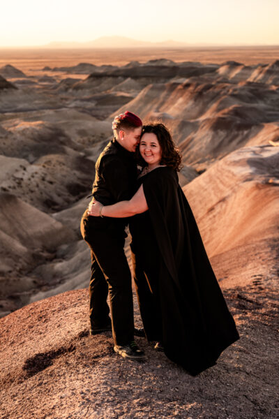 A couple in the Painted Desert sharing a quiet moment, one in a black wedding dress with a cape, standing between desert rocks.
