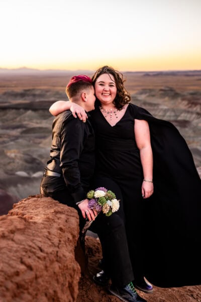 A queer couple in the Painted Desert, one bride in a black dress with a cape, embracing under the desert sky.