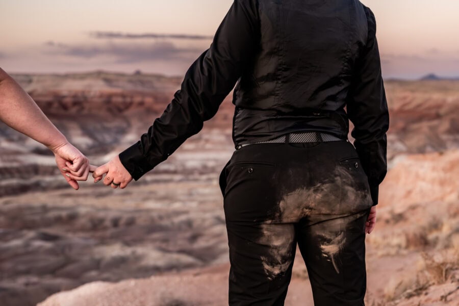 A queer couple in the Painted Desert, one in a black wedding dress with a cape, gazing at the desert landscape.