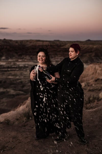 A queer couple and their friends toasting with champagne in the Painted Desert, one bride in a black wedding dress with a cape.