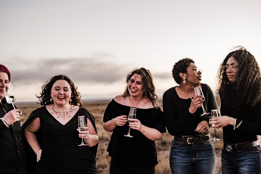 A queer couple and their friends celebrate with champagne in the Painted Desert, one bride in a black wedding dress with a cape.