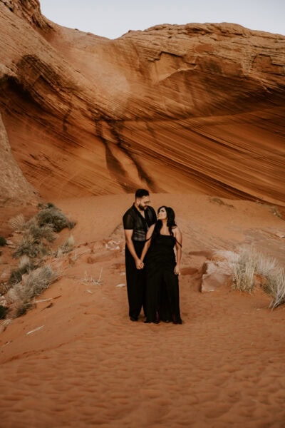 A man and woman in black wedding attire standing hand-in-hand in a red rock desert landscape, sharing a quiet moment.