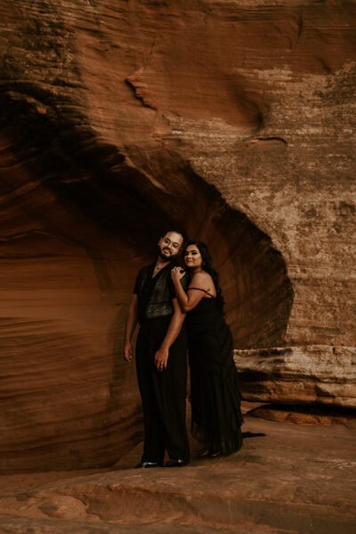 A couple in black wedding attire standing together against red rock cliffs, with the golden glow of sunset behind them