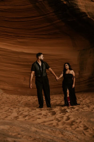 A man and woman in black attire walking hand-in-hand through a red rock desert, their silhouettes framed by the setting sun.