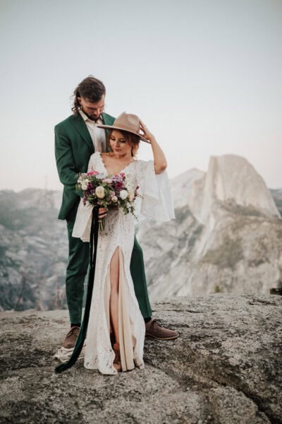 Elopement couple posing on the edge of a rocky overlook with Half Dome glowing at sunset